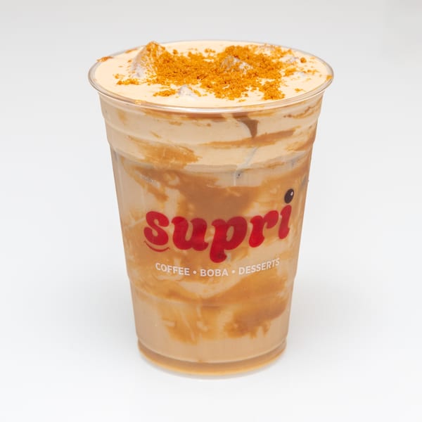 A Supri branded plastic cup filled with a caramel-colored drink, ice, and cookie crumbles.